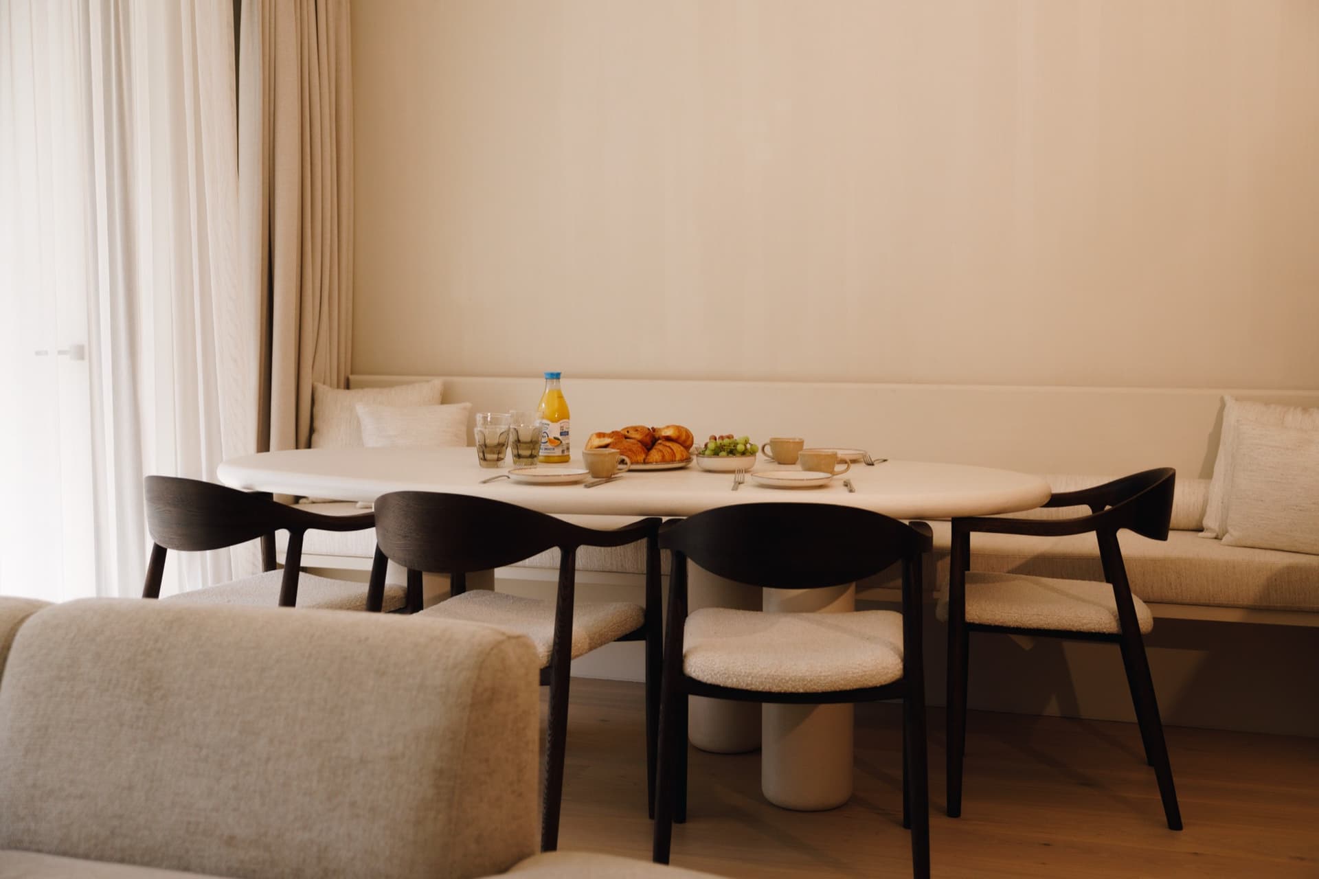 Ovale eettafel met vier donkere houten stoelen; ontbijt met croissants, sinaasappelsap en druiven, ingebouwde bank tegen de wand.