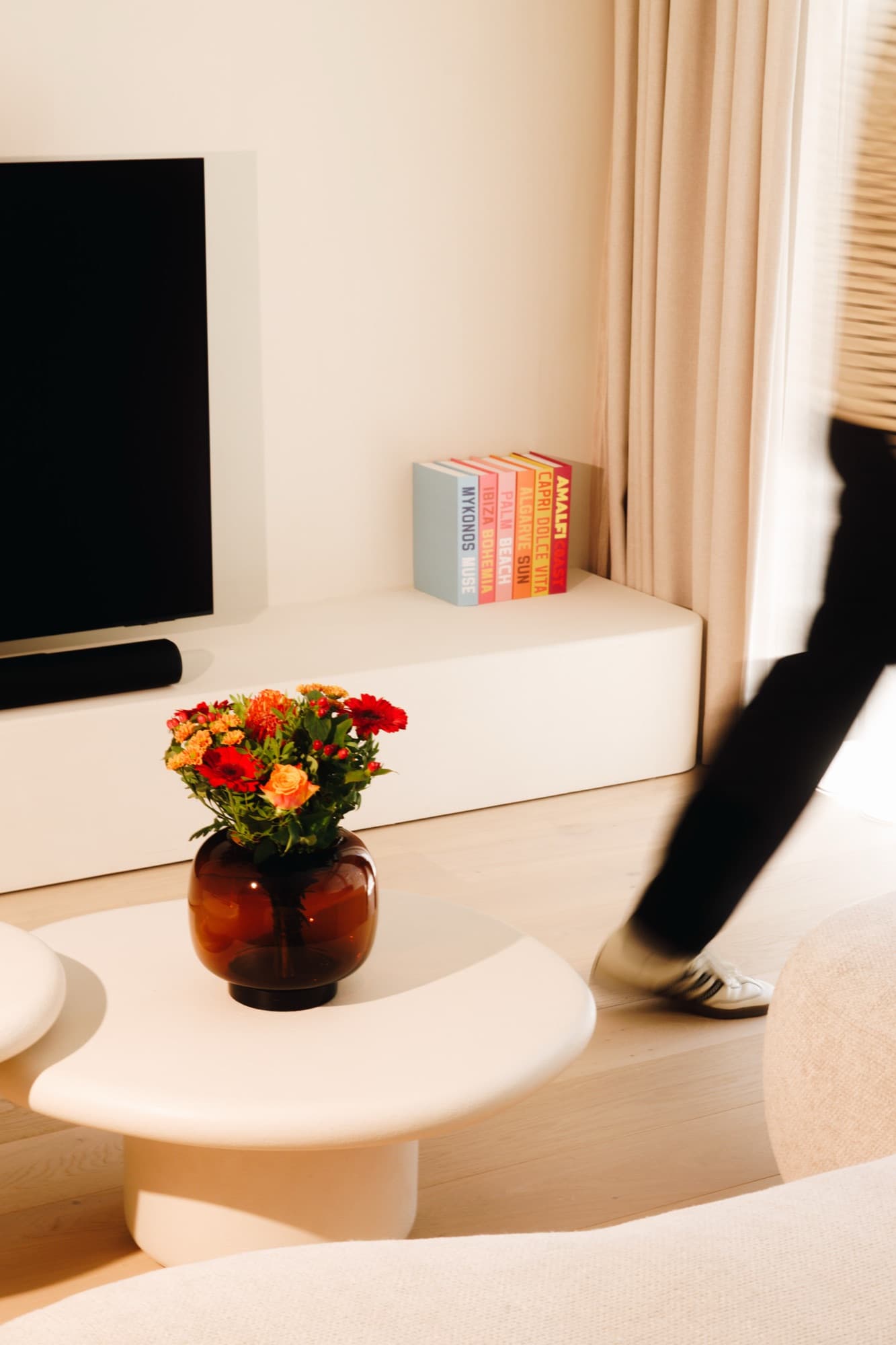 Lage witte salontafel met een amberkleurige vaas en herfstboeket; achter een tv-meubel met reisgidsen, iemand loopt voorbij.